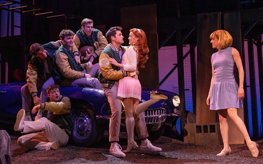 Stage scene from "The Outsiders" with characters interacting near a vintage car.