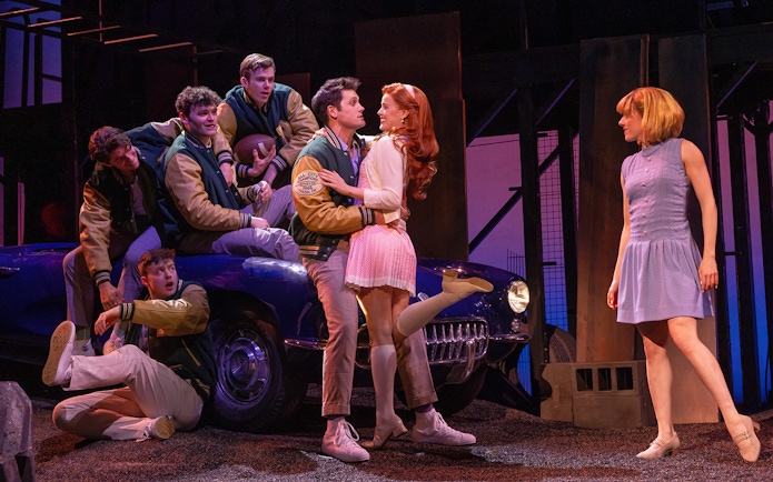 Stage scene from "The Outsiders" with characters interacting near a vintage car.