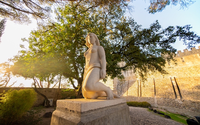 Statue of a woman at Sao Jorge Castle, Lisbon, with trees and castle wall in the background.