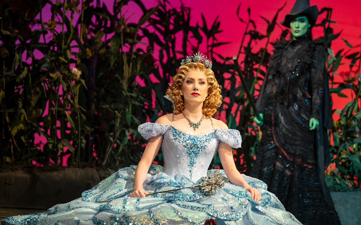 Blonde character in blue gown with wand, Wicked musical scene with green-skinned figure in background.