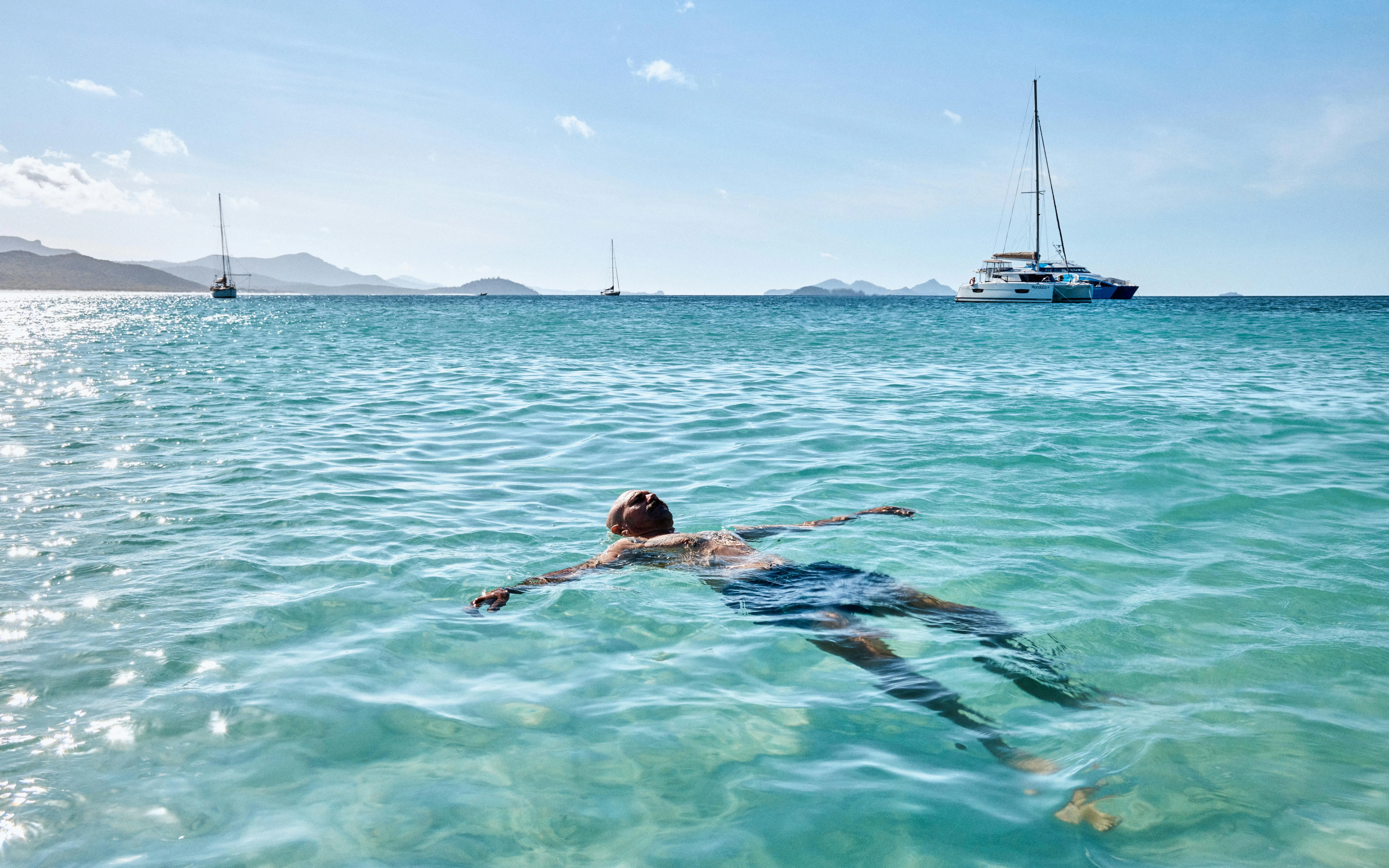 Person floating in clear water with sailboats in the distance at Whitehaven Beach, Whitsundays.