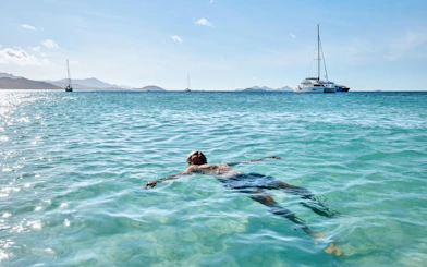 Person floating in clear water with sailboats in the distance at Whitehaven Beach, Whitsundays.