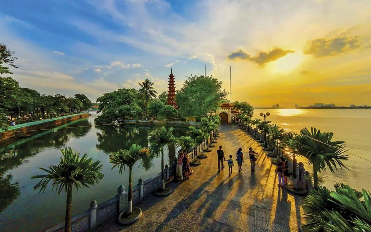 Tran Quoc Pagoda at sunset on West Lake, Hanoi, with visitors walking along the palm-lined path.