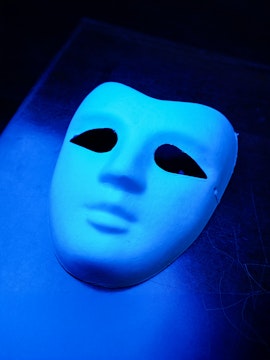 Blue theatrical mask with black eye holes on a dark background.