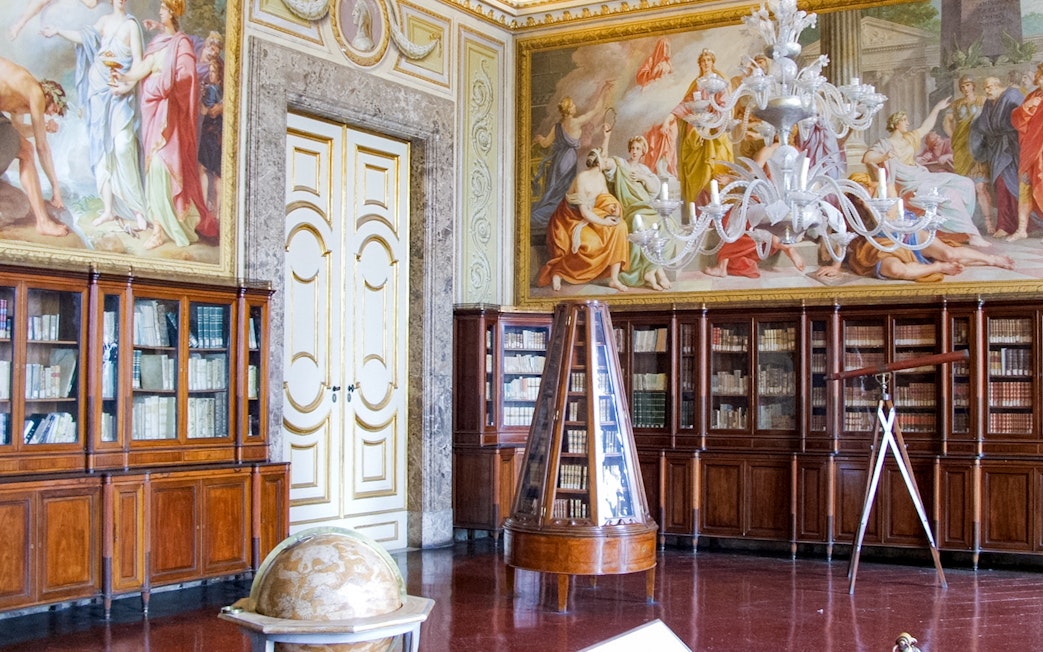 Royal Palace of Caserta library with ornate ceiling frescoes and wooden bookcases.