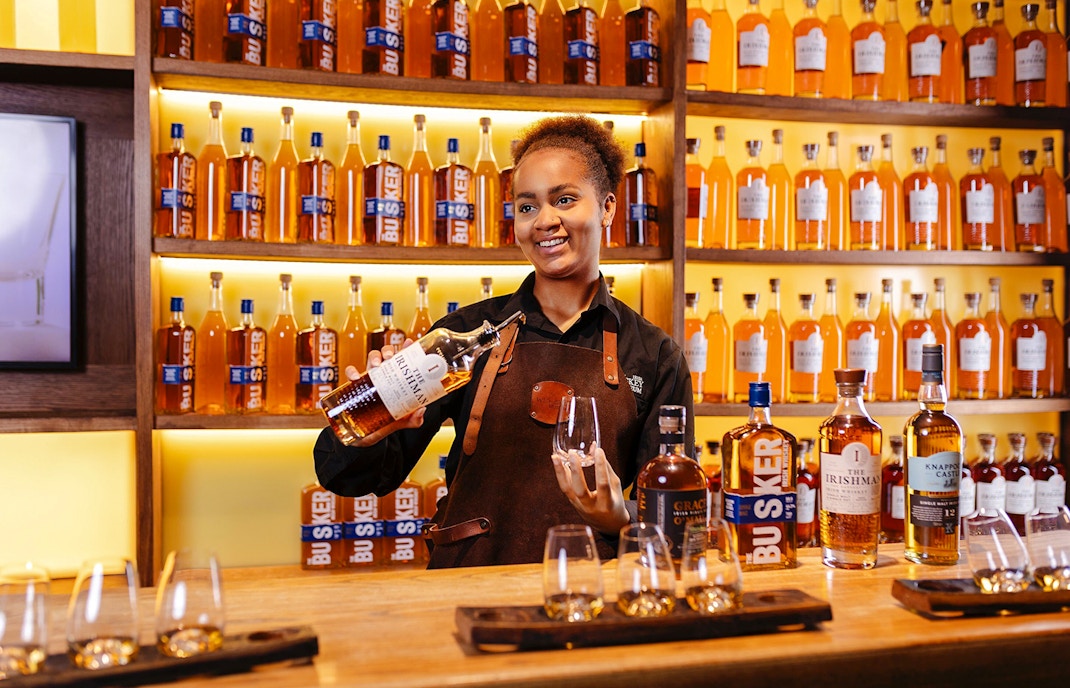 Host pouring whiskey at the Irish Whiskey Museum with bottles displayed in the background.