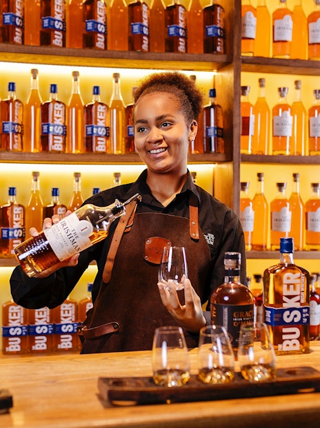 Host pouring whiskey at the Irish Whiskey Museum with bottles displayed in the background.