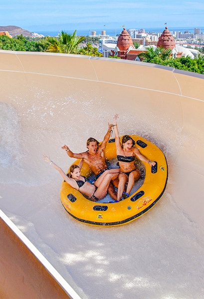 Group enjoying a water ride at Siam Park, Tenerife, with cityscape in the background.