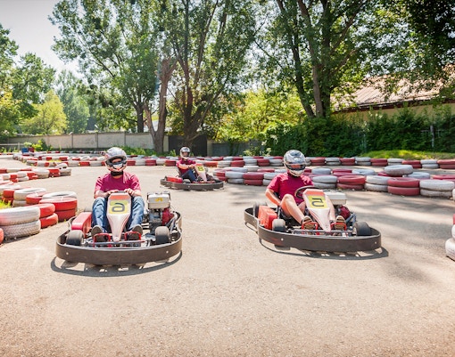 Three people racing go-karts on an outdoor circuit surrounded by trees and tire barriers.