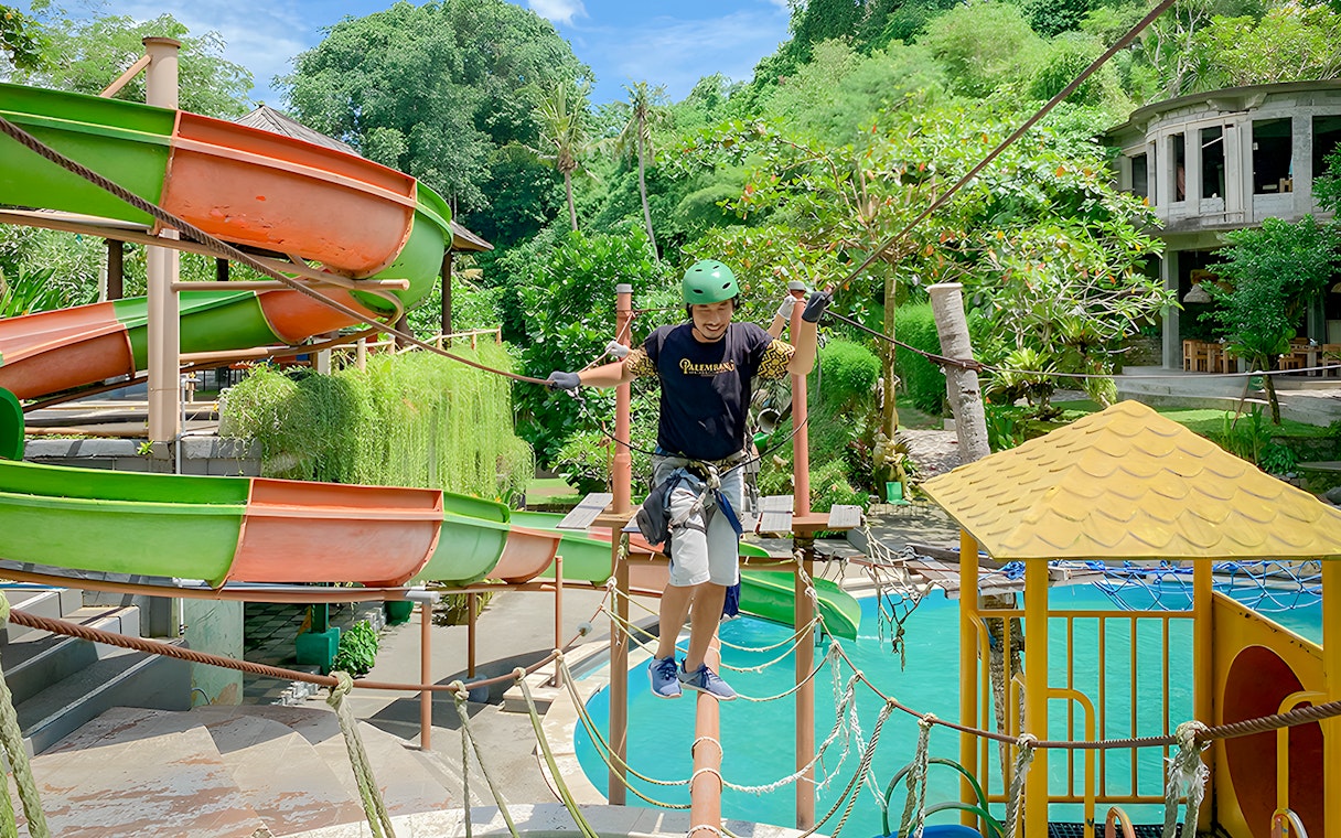 Person crossing a rope bridge at Keramas Park with water slides in the background.