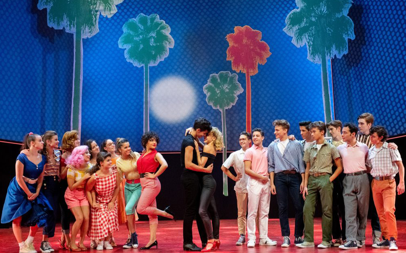 Cast of Grease performing on stage with colorful palm tree backdrop.