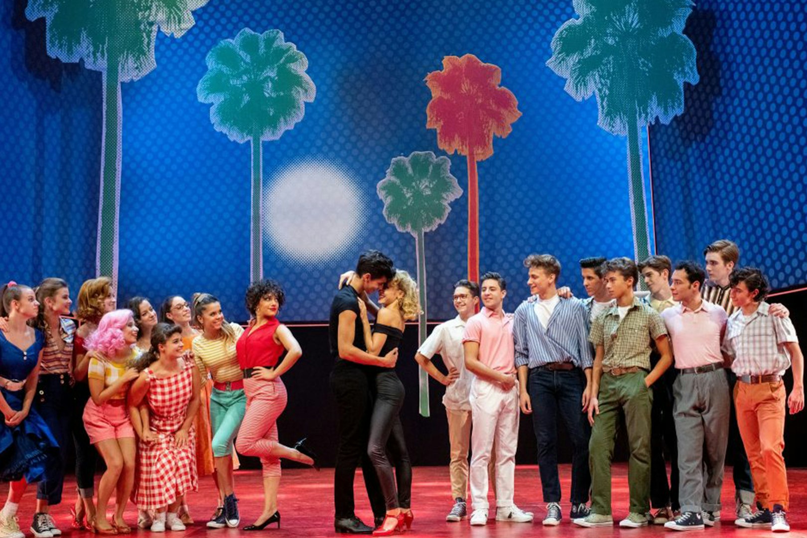 Cast of Grease performing on stage with colorful palm tree backdrop.