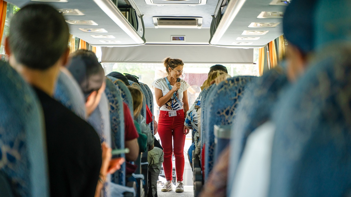 Tour guide speaking to passengers on a bus during a guided tour transfer.