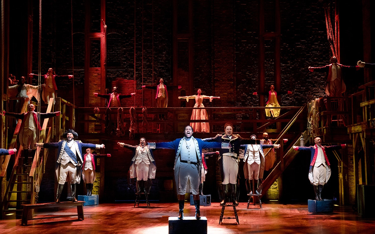 Stage performance of Hamilton cast in period costumes with arms outstretched.