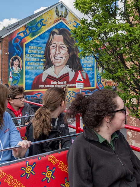 Tourists on Belfast hop-on-hop-off bus viewing mural of Bobby Sands on city street.