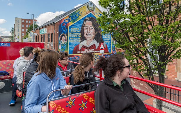 Tourists on Belfast hop-on-hop-off bus viewing mural of Bobby Sands on city street.
