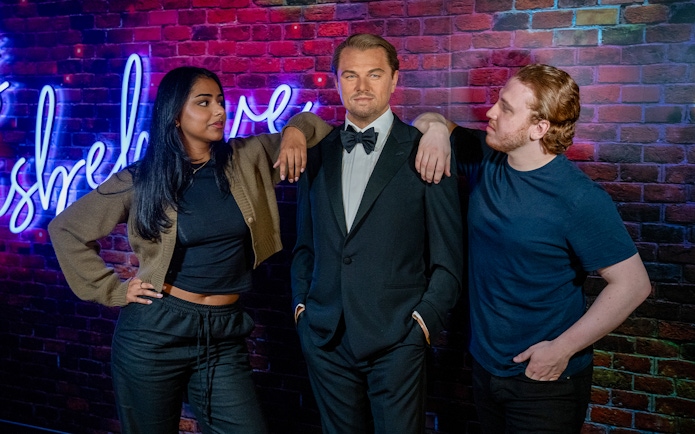 Wax figure of a man in a tuxedo at Madame Tussauds, London, with two visitors posing beside it.