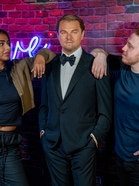 Wax figure of a man in a tuxedo at Madame Tussauds, London, with two visitors posing beside it.