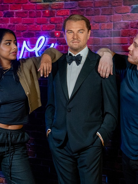 Wax figure of a man in a tuxedo at Madame Tussauds, London, with two visitors posing beside it.
