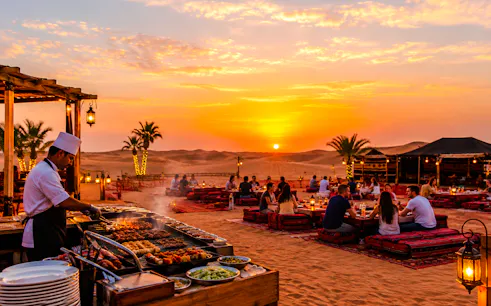 Chef grilling food at sunset during Dubai Elite Red Dune Safari BBQ, with guests dining on desert cushions.