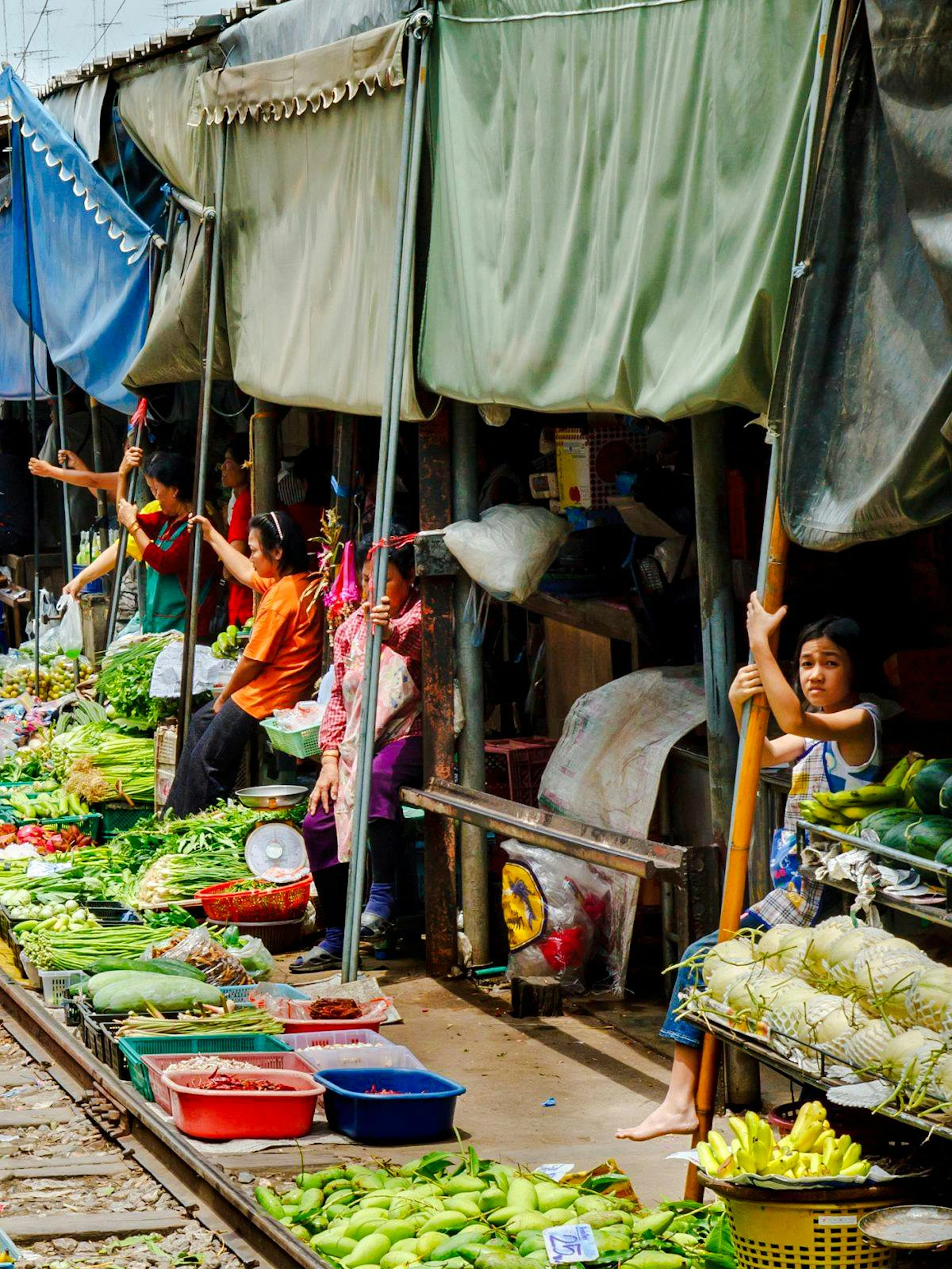 Train passing through Maeklong Railway Market with vendors and produce on both sides.