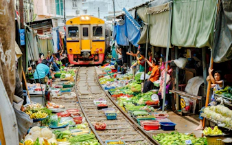 Train passing through Maeklong Railway Market with vendors and produce on both sides.