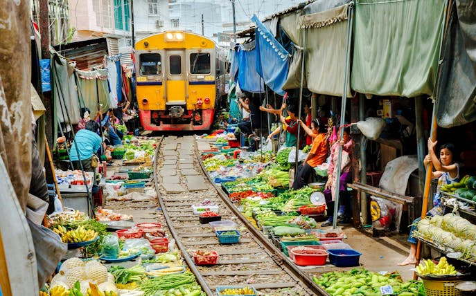 Train passing through Maeklong Railway Market with vendors and produce on both sides.