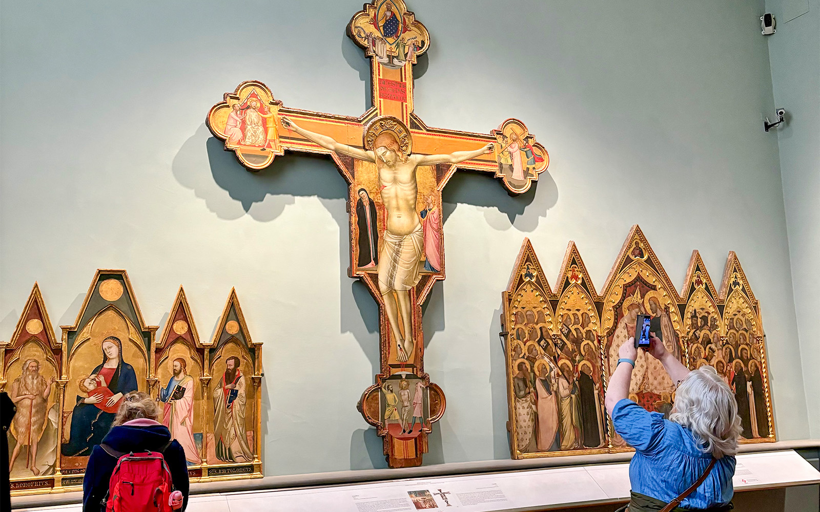 Crucifix and altarpieces at Accademia Art Gallery, Florence, with visitors observing.