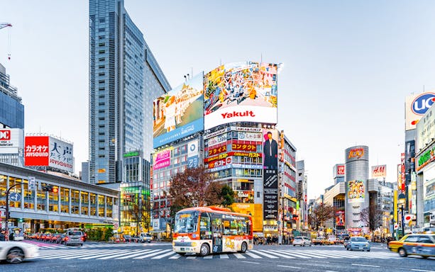 Shibuya Crossing in Tokyo with vibrant billboards and a tour bus.