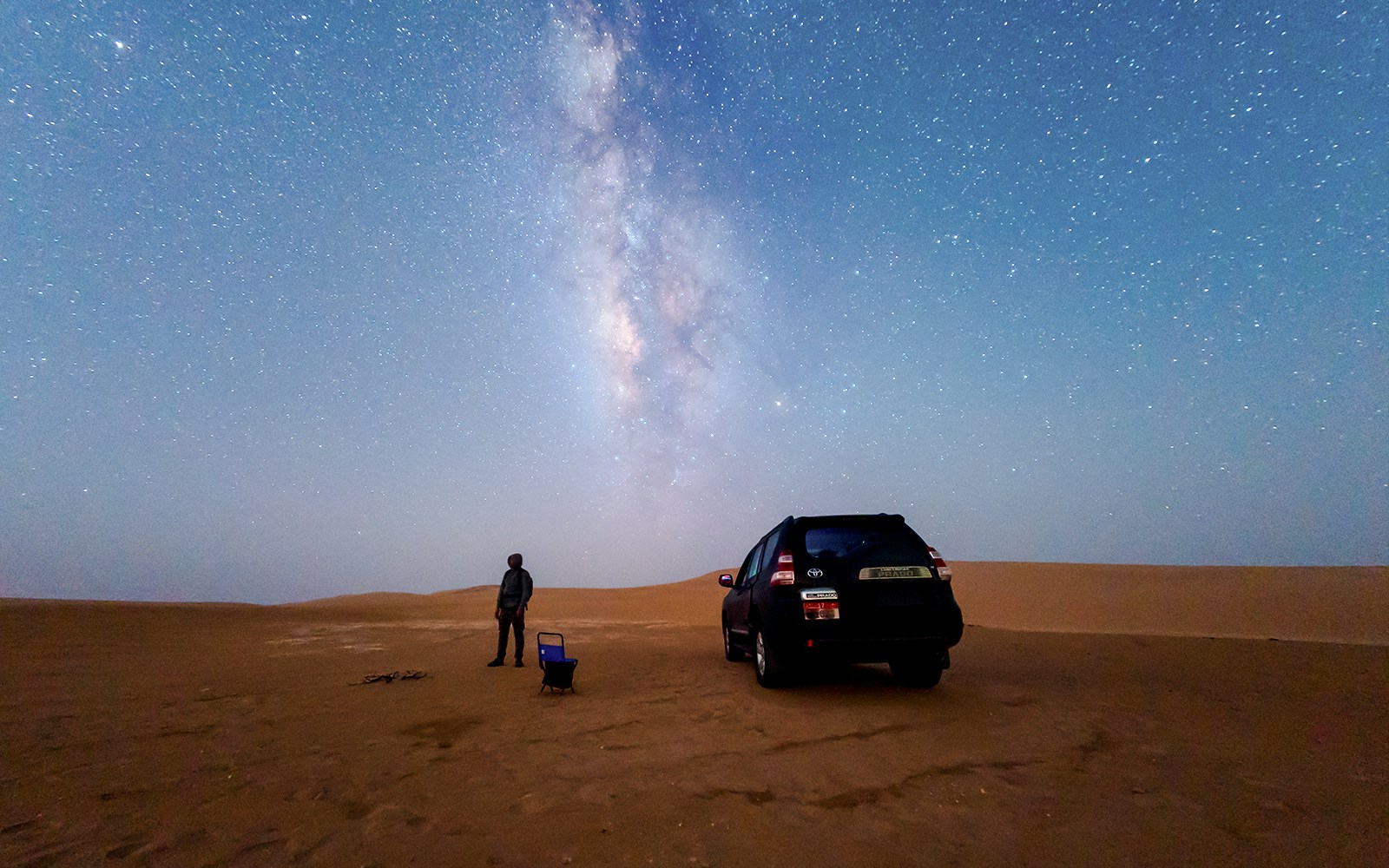 Milky Way over desert sand dune with person and parked car.