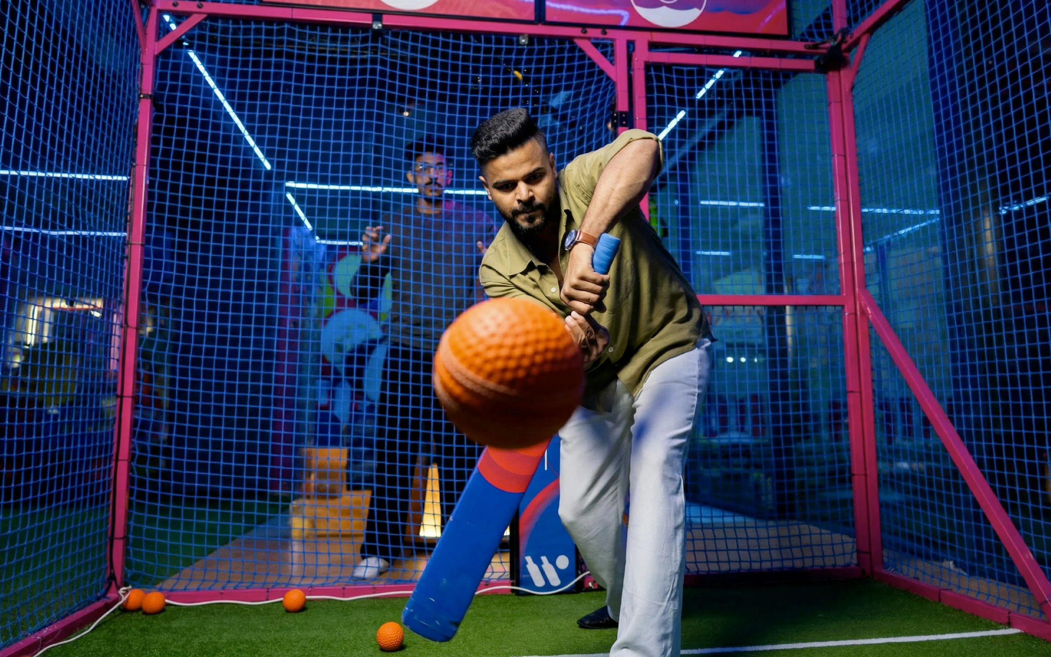 Man playing cricket in an indoor batting cage with a blue bat and orange ball.