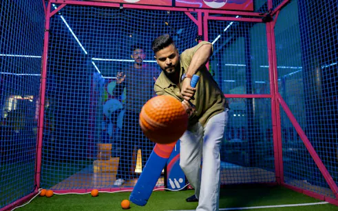 Man playing cricket in an indoor batting cage with a blue bat and orange ball.