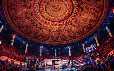 Boxing match at Rajadamnern Stadium under ornate ceiling.