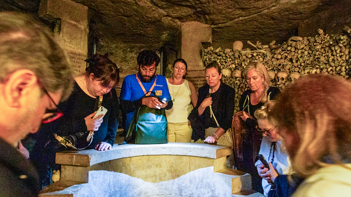 Tourists exploring the Paris Catacombs, viewing skulls and bones in dimly lit tunnels.