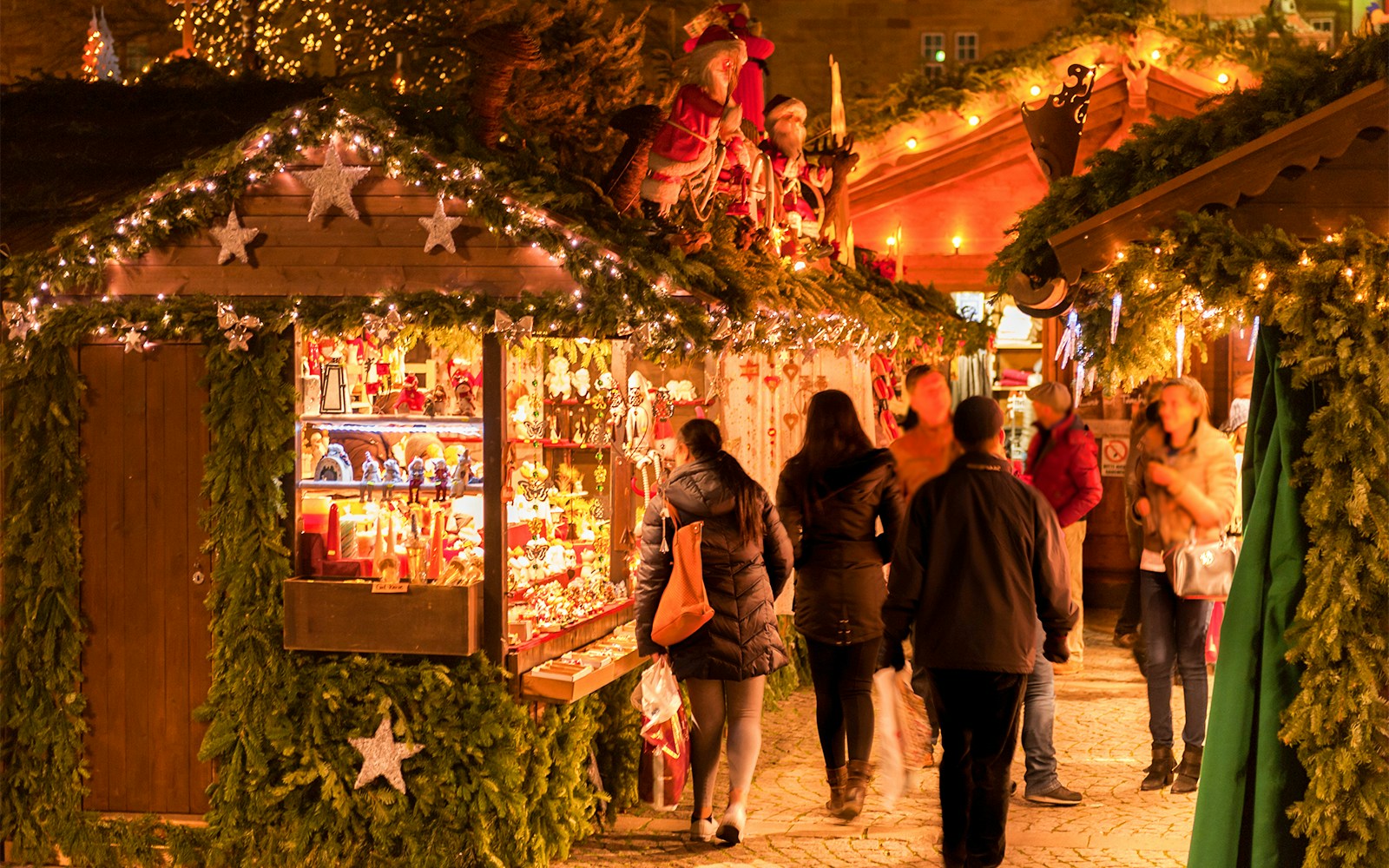 Christmas market stalls with festive lights and decorations, people browsing in the evening.