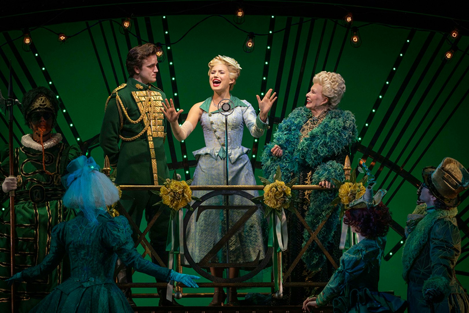 Cast performing a scene from the musical Wicked on stage with green backdrop.