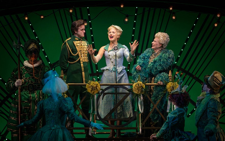 Cast performing a scene from the musical Wicked on stage with green backdrop.
