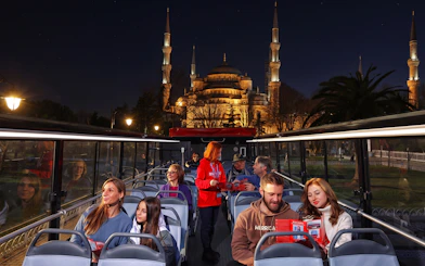 Open-top bus tour in Istanbul with view of illuminated Blue Mosque at night.