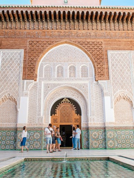 Visitors at the ornate entrance of Madrasa Ben Youssef, Marrakech, with intricate tilework and carvings.