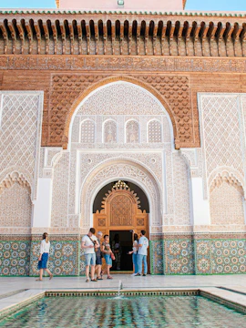 Visitors at the ornate entrance of Madrasa Ben Youssef, Marrakech, with intricate tilework and carvings.