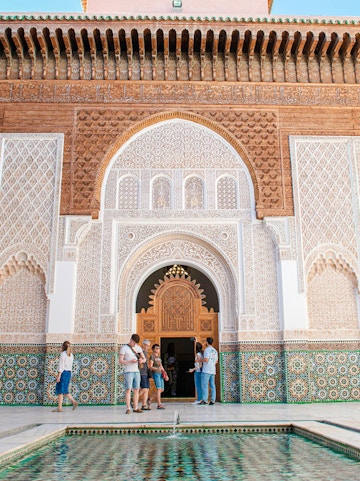 Visitors at the ornate entrance of Madrasa Ben Youssef, Marrakech, with intricate tilework and carvings.