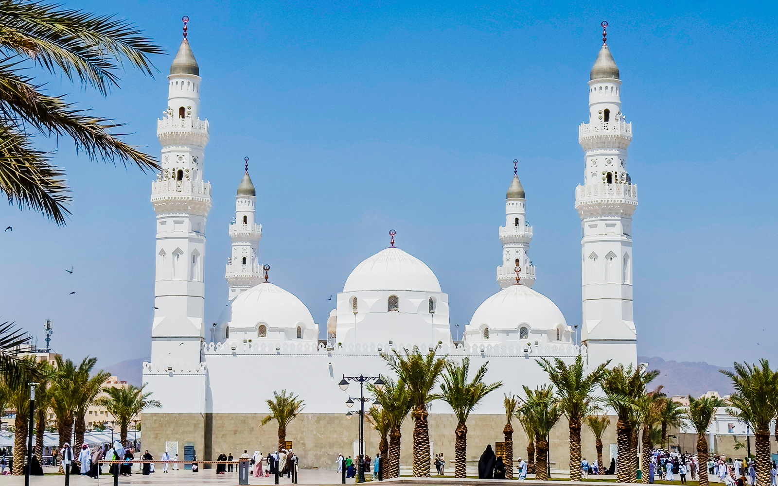 Quba Mosque in Medina, Saudi Arabia, with palm trees and visitors in the courtyard.