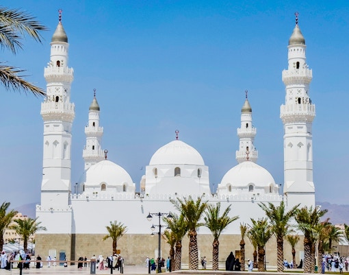 Quba Mosque in Medina, Saudi Arabia, with palm trees and visitors in the courtyard.