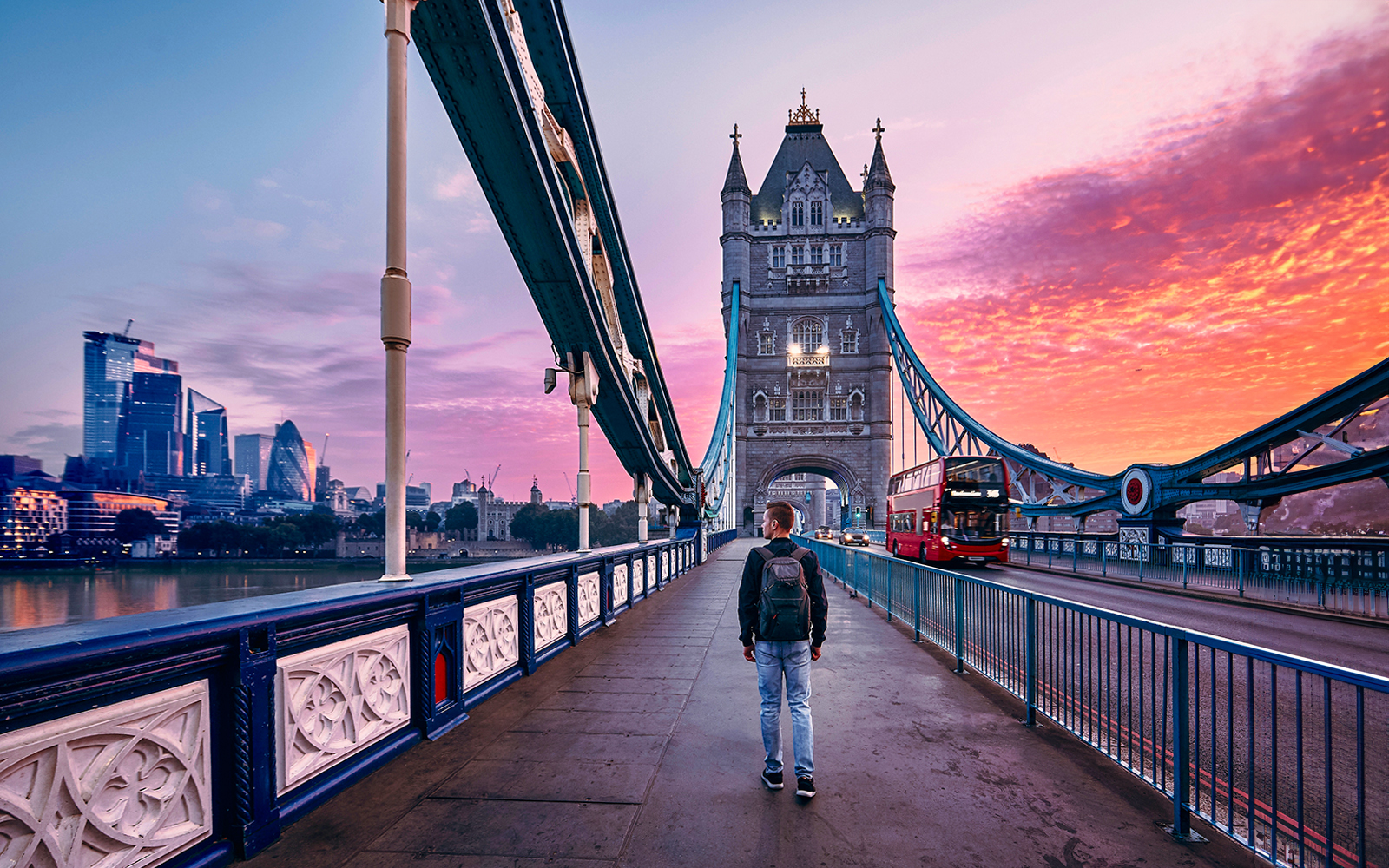 Person walking on Tower Bridge in London at sunset with a red bus passing by.