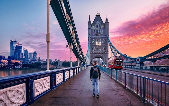Person walking on Tower Bridge in London at sunset with a red bus passing by.