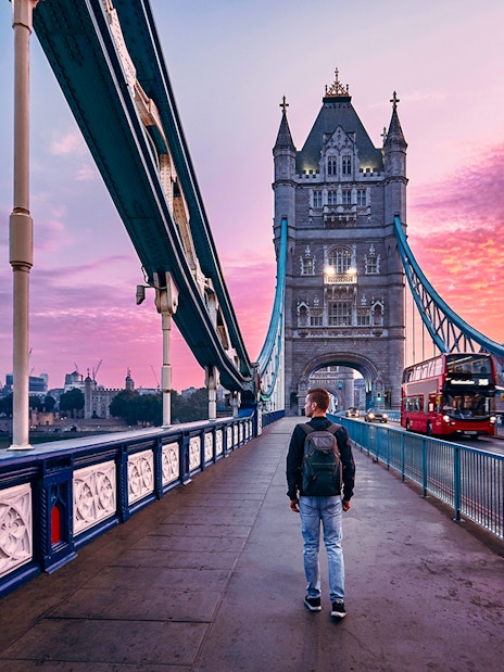 Person walking on Tower Bridge in London at sunset with a red bus passing by.