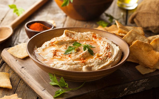 Hummus garnished with paprika and parsley, served with pita chips on a wooden board.