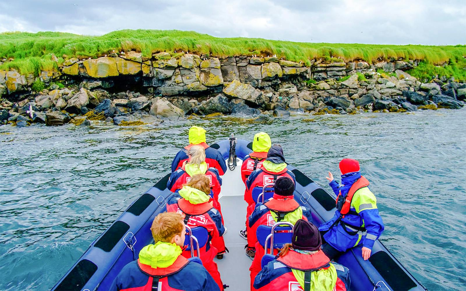 RIB boat with passengers in red jackets approaching rocky coastline for whale watching.