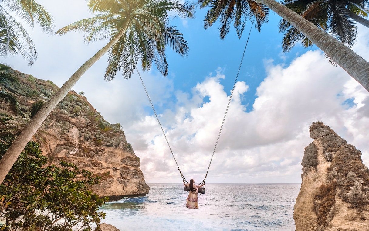 Person on a swing between palm trees overlooking ocean cliffs, Nusa Penida Island.