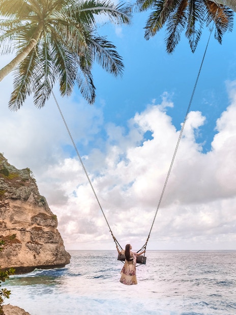Person on a swing between palm trees overlooking ocean cliffs, Nusa Penida Island.